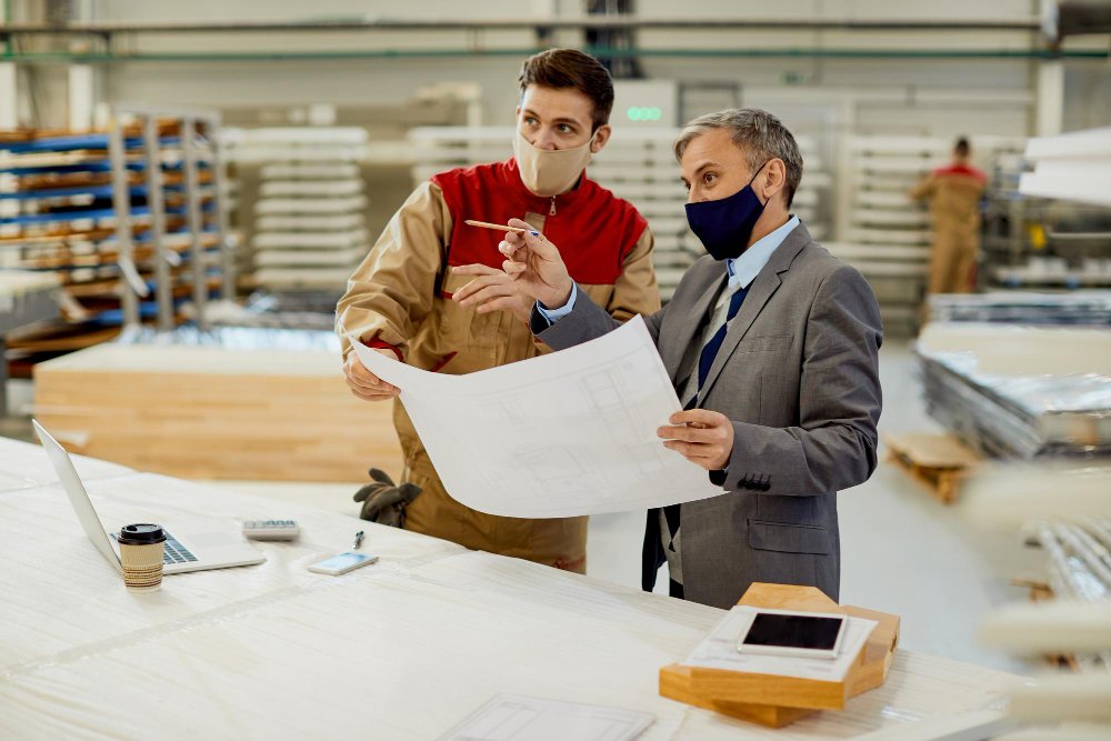 Two men wearing suits and masks are engaged in work at a factory, showcasing a commitment to safety in artisan contracting.